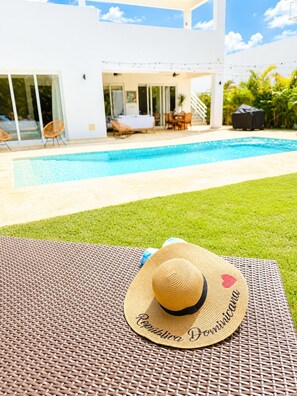 A poolside area with a lounge chair, a sun hat, and a view of a modern house with a pool. - 3bd with rooftop ocean view in Sosua Ocean Village (Sosúa)