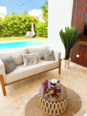 A modern outdoor seating area with a sofa, a round wooden table with drinks, and a view of a swimming pool. - 3bd with rooftop ocean view in Sosua Ocean Village (Sosúa)