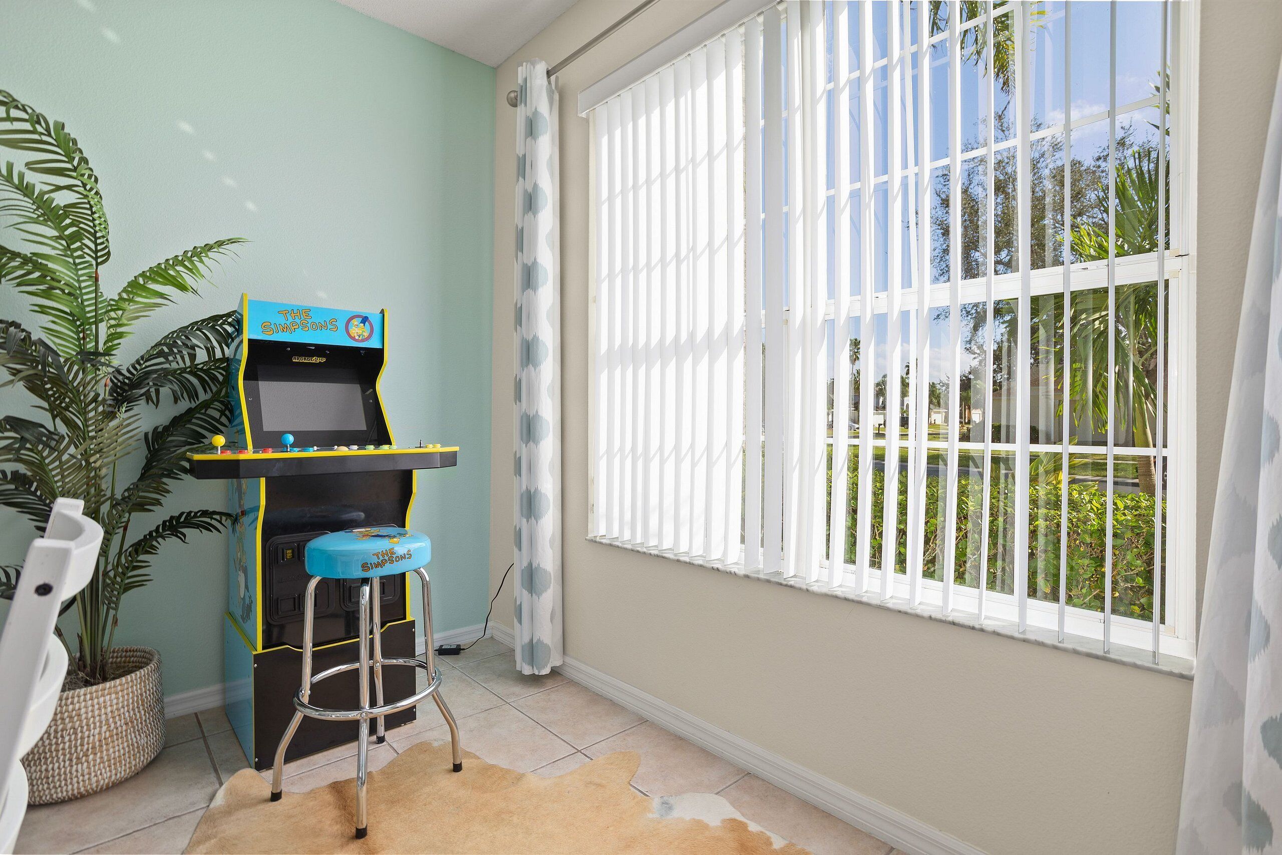 A room with a play area featuring a blue arcade machine, a large potted plant, and a window with white blinds.