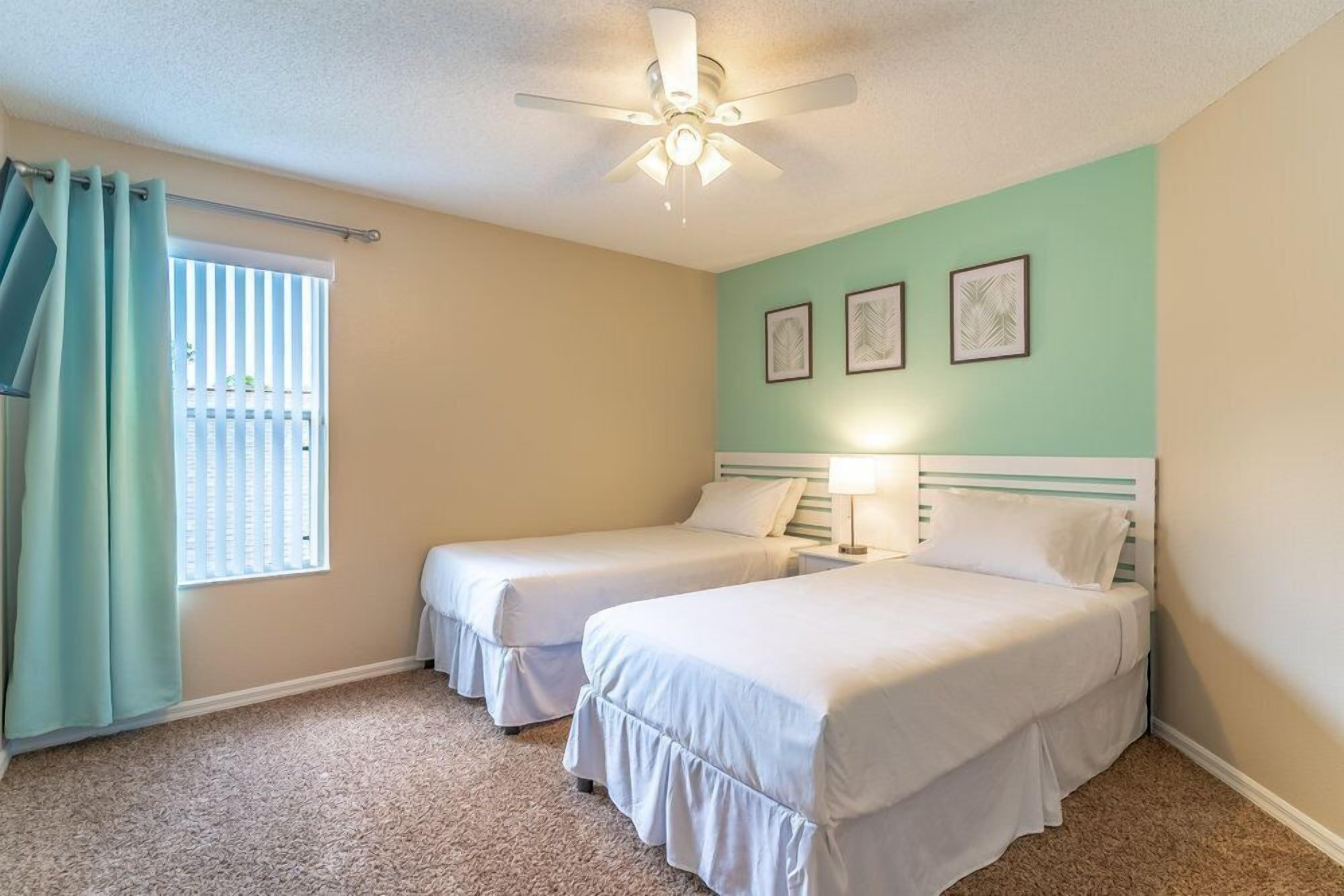 A room with two single beds, a ceiling fan, a window with curtains, and three framed pictures on the wall.
