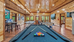 A pool table with colorful balls, a billiards table, and a ceiling fan in a wooden cabin with large windows. - Private Indoor Heated Pool, Arcade, HotTub Sleeps24 (Sevierville)