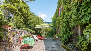 Un patio empedrado con una mesa y sillas, una sombrilla verde y abundante vegetación.