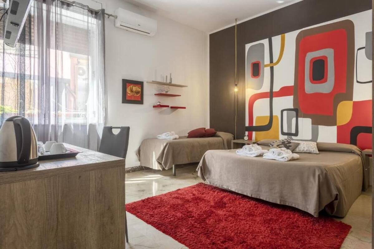 Una habitació d'hotel amb dos llits, una catifa vermella, una taula de nit amb un bullidor i un quadre abstracte gran a la paret.