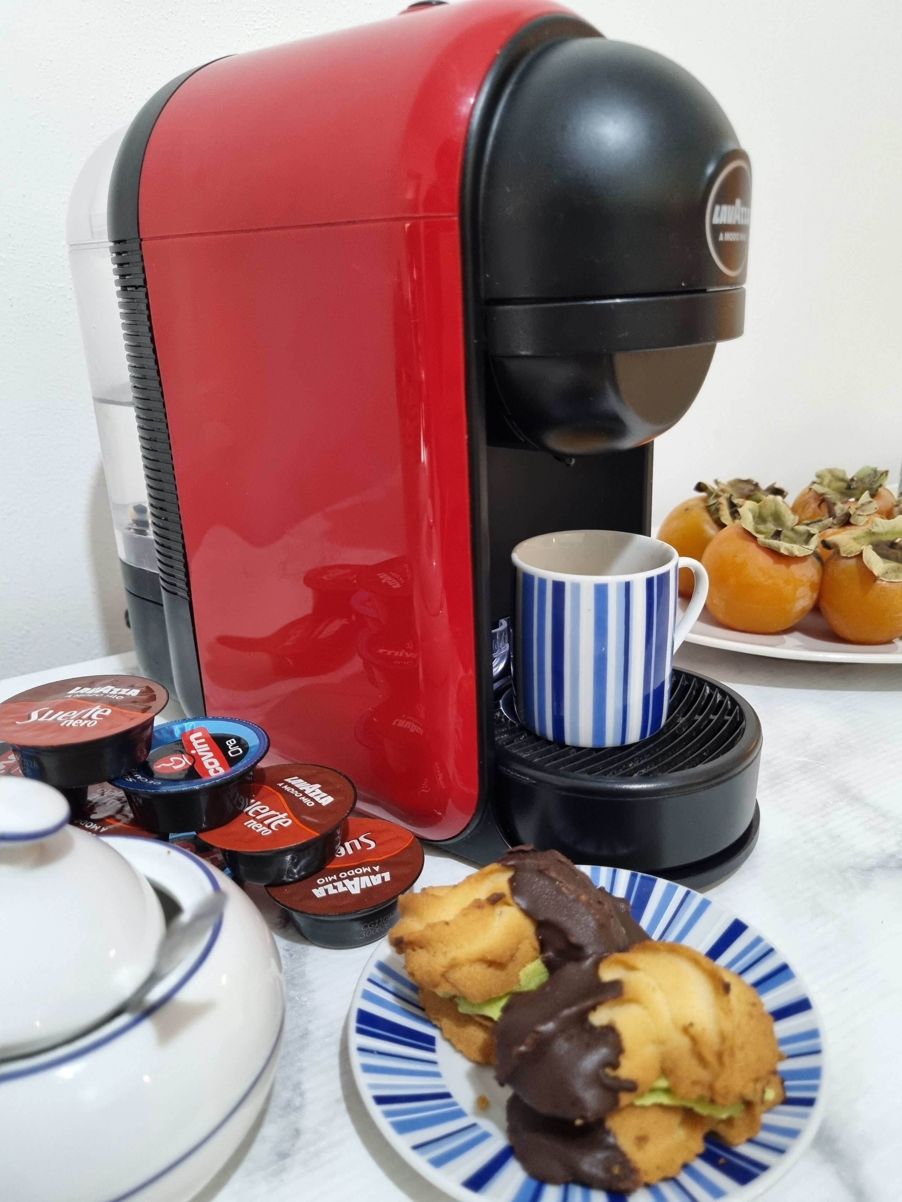 A red and black coffee maker, a blue and white striped mug, and a plate with pastries and coffee pods.