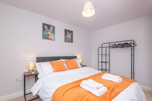 A neatly made bed with white and orange bedding, two wall-mounted paintings, a hanging light fixture, and a metal rack with clothes.
