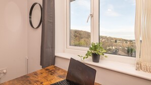 Una habitación con una computadora portátil sobre una mesa de madera, una planta en maceta en el alféizar de la ventana, un aro en la pared y vistas a un pueblo a través del ventanal.