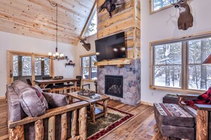 L’intérieur chaleureux d’un chalet avec une cheminée en pierre, des meubles en bois et de grandes fenêtres donnant sur le paysage enneigé.