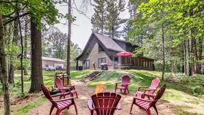 Uma cabana com lareira cercada por cadeiras Adirondack vermelhas, situada numa área arborizada com um caminho de terra que leva à cabana.