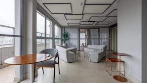 A modern living room with a sofa, chairs, a coffee table, and a small side table. There are large windows with curtains, a potted plant, and a view of the city. - Stay Steps fr/ the Metro and Ibirapuera Mall in Indianopolis - Noes (São Paulo)