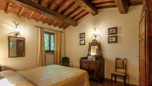A bedroom with a wooden ceiling, a bed with a beige bedspread, a wooden dresser, a chair, and a mirror with framed pictures on the wall. - Vin Santo – Independence and Relaxation with Private Garden (Reggello)
