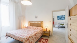 A bedroom with a bed, bedside lamps, a nightstand, a wooden dresser, and a view into another room. - VILLA LA SCIROCCATA LEUCA (Santa Maria di Leuca)