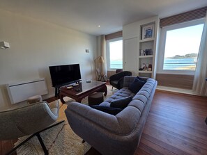 Sala de estar con un sofá, televisor, mesa de centro y una silla. Se ve una ventana con vista al mar.