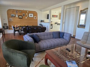Una sala de estar moderna con un sofá gris, un sillón verde, una mesa de centro de vidrio y una pared de madera con cuadros enmarcados.