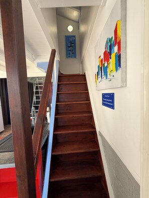 Eine hölzerne Treppe führt zu einer oberen Etage, an der Wand ist ein blaues Schild und darüber ein farbenfrohes abstraktes Gemälde angebracht.