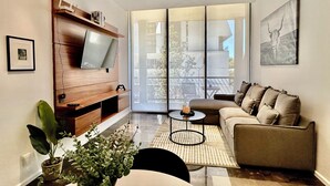 A modern living room with a flat-screen TV mounted on a wooden shelf, a sofa, a coffee table, and a large sliding glass door leading to an outdoor area. - El DePA, Luxury and Comfort, resort-like amenities (Zapopan)