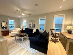 A living room with a dark blue sofa, a white armchair, a wooden coffee table, and a wooden chair. There is a ceiling fan, a painting on the wall, and a window with blinds.