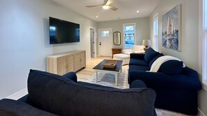A modern living room with a large flat-screen TV, dark blue sofas, a wooden cabinet, a ceiling fan, and a painting on the wall. - Cozy Cottage Retreat • 5 Min to Cedar Point • Deck + Screened Patio (Sandusky)