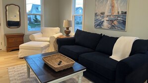 A living room with a dark blue sofa, a white armchair, a wooden coffee table with a woven basket, a lamp, and a painting of sailboats. - Cozy Cottage Retreat • 5 Min to Cedar Point • Deck + Screened Patio (Sandusky)