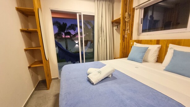 A hotel room with a large bed, a wooden shelf, a window with curtains, and a view of palm trees. - Chalet with private garden, ideal for couples and families with children. (São Miguel do Gostoso)