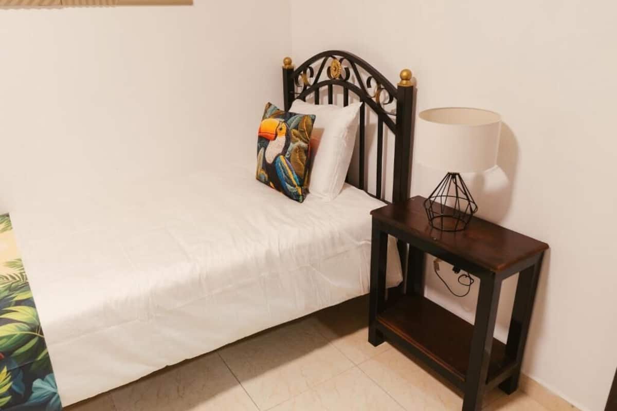 Un lit avec un drap blanc, une tête de lit en métal noir et un oreiller coloré. Une petite table de chevet en bois, sur laquelle se trouvent une lampe et un objet décoratif, est placée à côté du lit.