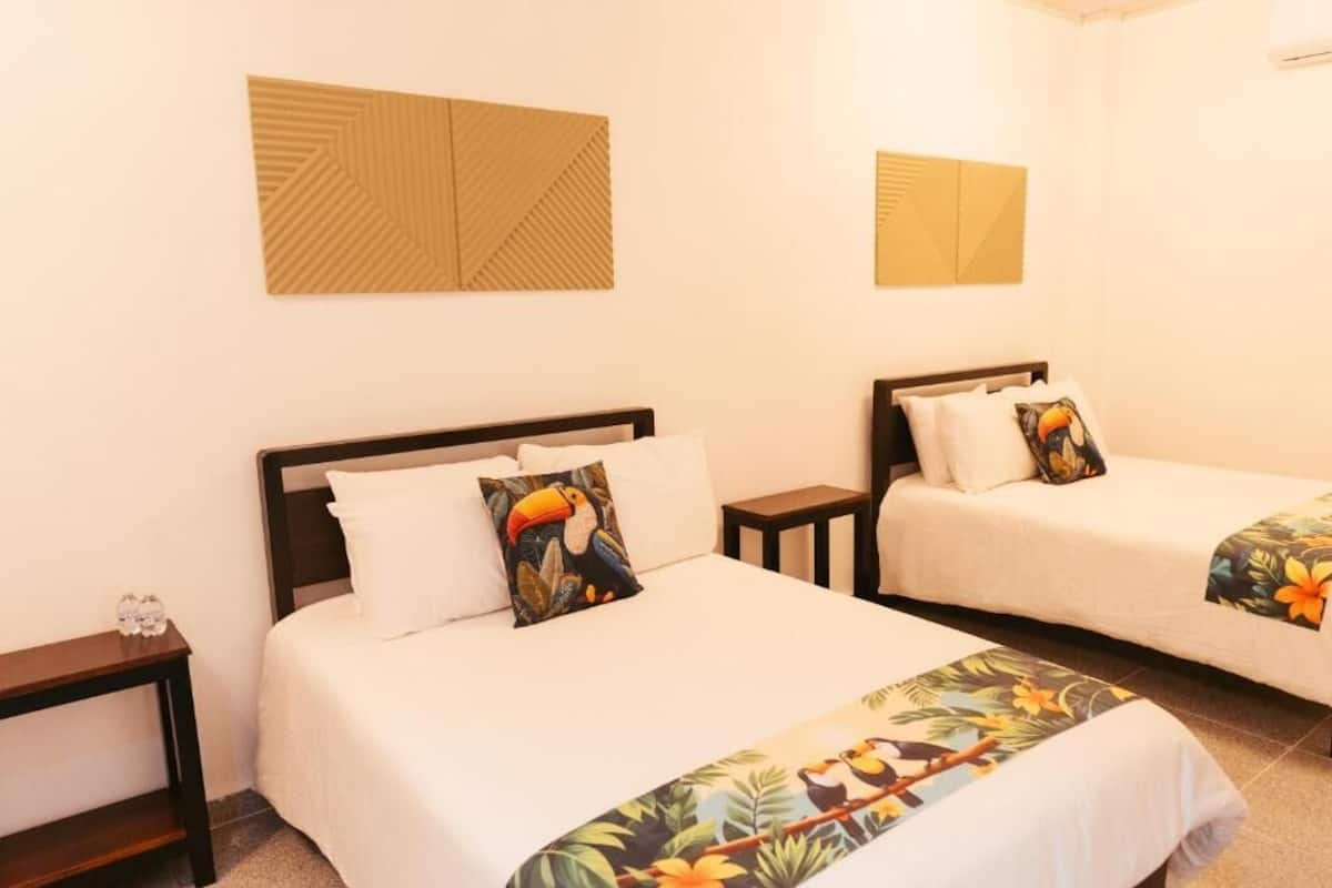 Une chambre d’hôtel avec deux lits, chacun recouvert d’une courtepointe blanche et agrémenté d’un coussin aux motifs tropicaux. Il y a deux tables de chevet, une de chaque côté des lits, et un miroir fixé au mur au-dessus des lits.