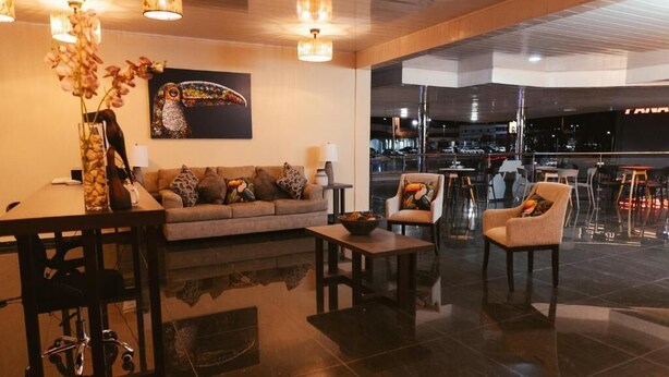 A modern lobby with a sofa, armchairs, a desk, and a large window offering a view of the cityscape. - Hotel Plaza Los Tucanes (Nuevo Santiago)