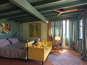 A room with a bed, a yellow sofa, a round table with a lamp, a painting on the wall, and a window with curtains. - Kalliberry Cycladic hidden gem with outdoor bathtub & amazing sunsets (Andros)