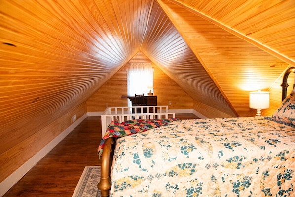 Ein gemütliches Schlafzimmer im Dachgeschoss mit Holzbalken an der Decke, einem Bett mit floralem Bettzeug, einem Nachttisch und einem kleinen Tisch mit Lampe.