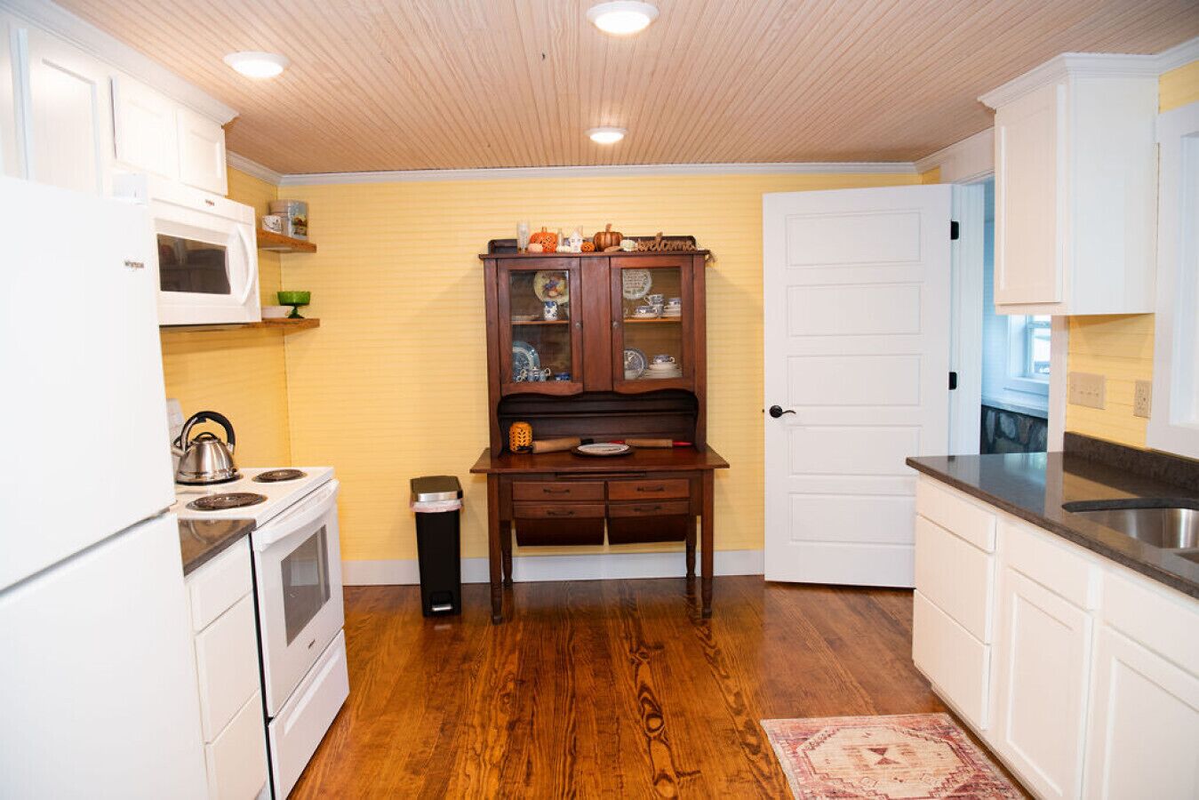 A compact kitchen with white cabinets, a wooden floor, a microwave, a stove, and a wooden cabinet with dishes.