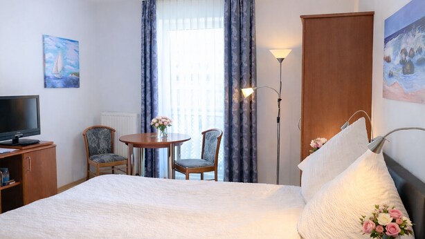 Ett hotellrum med en säng, ett skrivbord med en tv, två stolar, ett bord med blommor, en lampa och konstverk på väggarna.