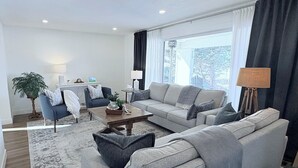 En moderne stue med en grå sofa, et sofabord og et lille bord med en lampe. Der er en potteplante og et lille bord med pyngeting.