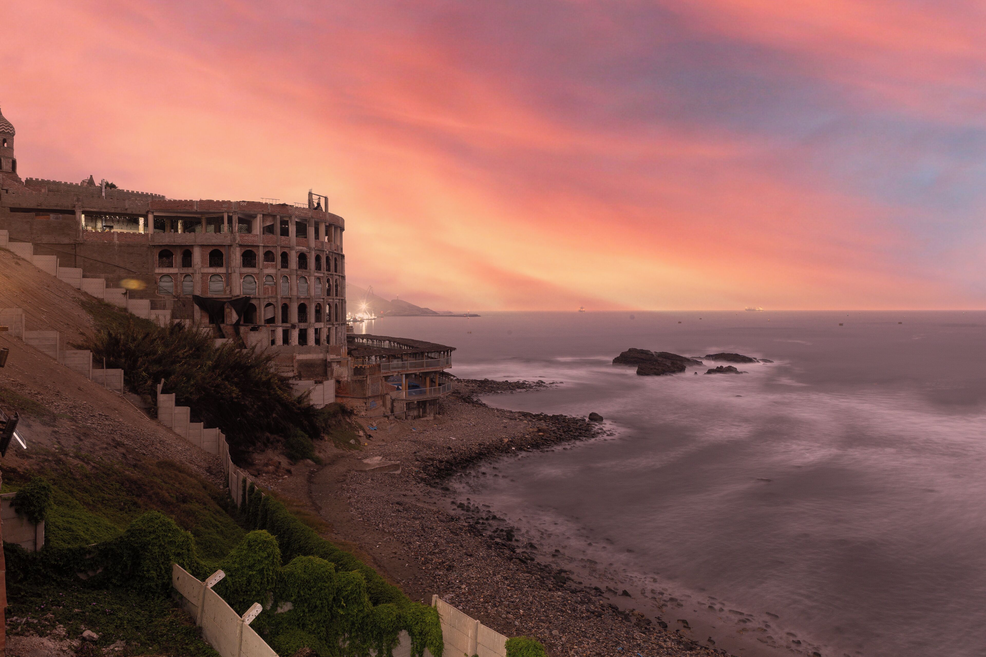A coastal building with multiple balconies overlooking the sea, a staircase leading up to the building, and a rocky shoreline.