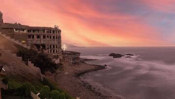 A coastal building with multiple balconies overlooking the sea, a staircase leading up to the building, and a rocky shoreline.