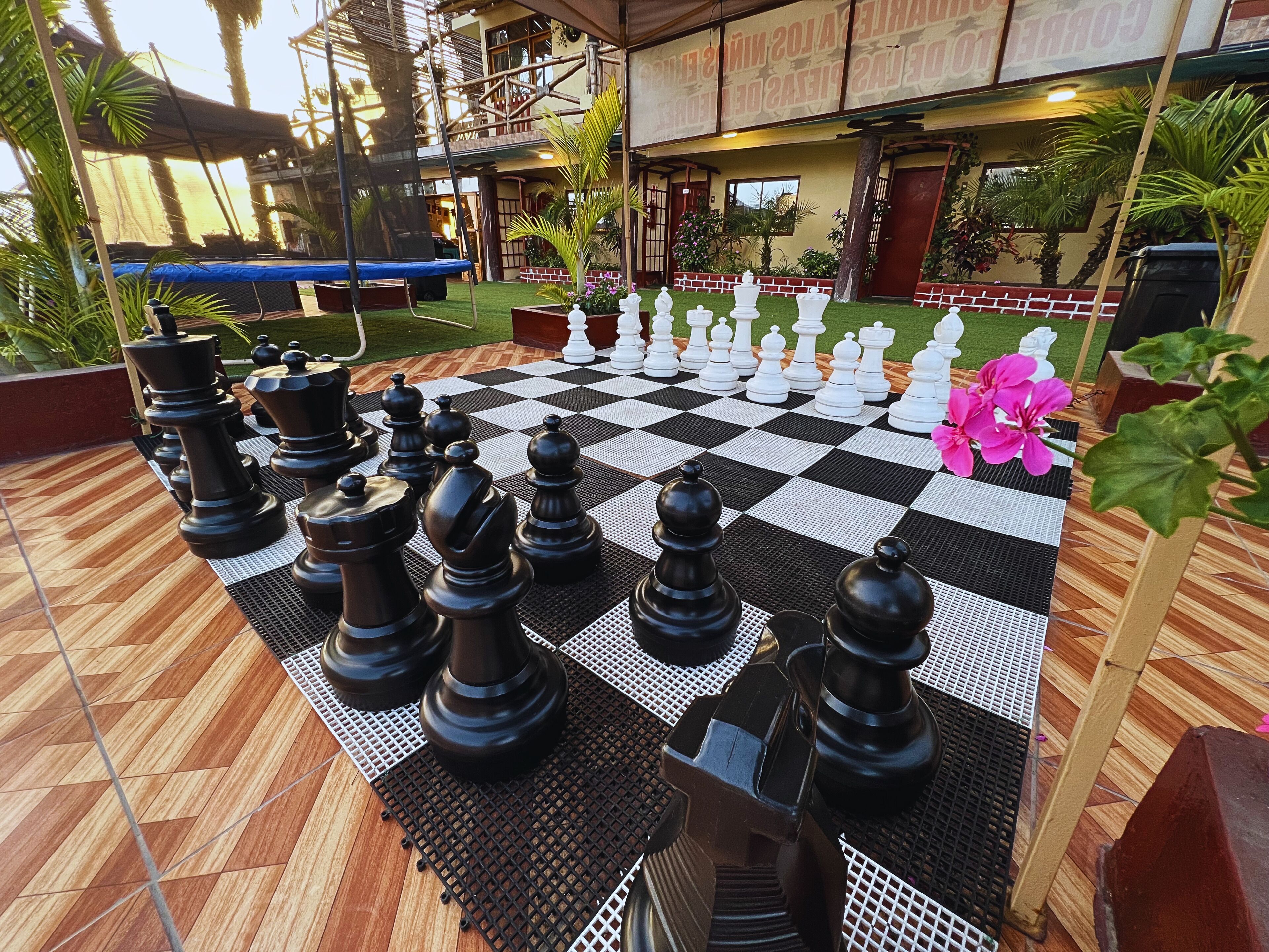 Un jeu d’échecs installé en plein air, sur un sol à carreaux, entouré de verdure et avec un bâtiment en arrière-plan.