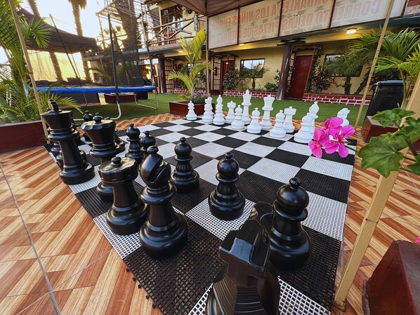 A chessboard set outdoors with a checkered floor, surrounded by greenery and a building in the background. - Hotel Sunset Chancay (Chancay)