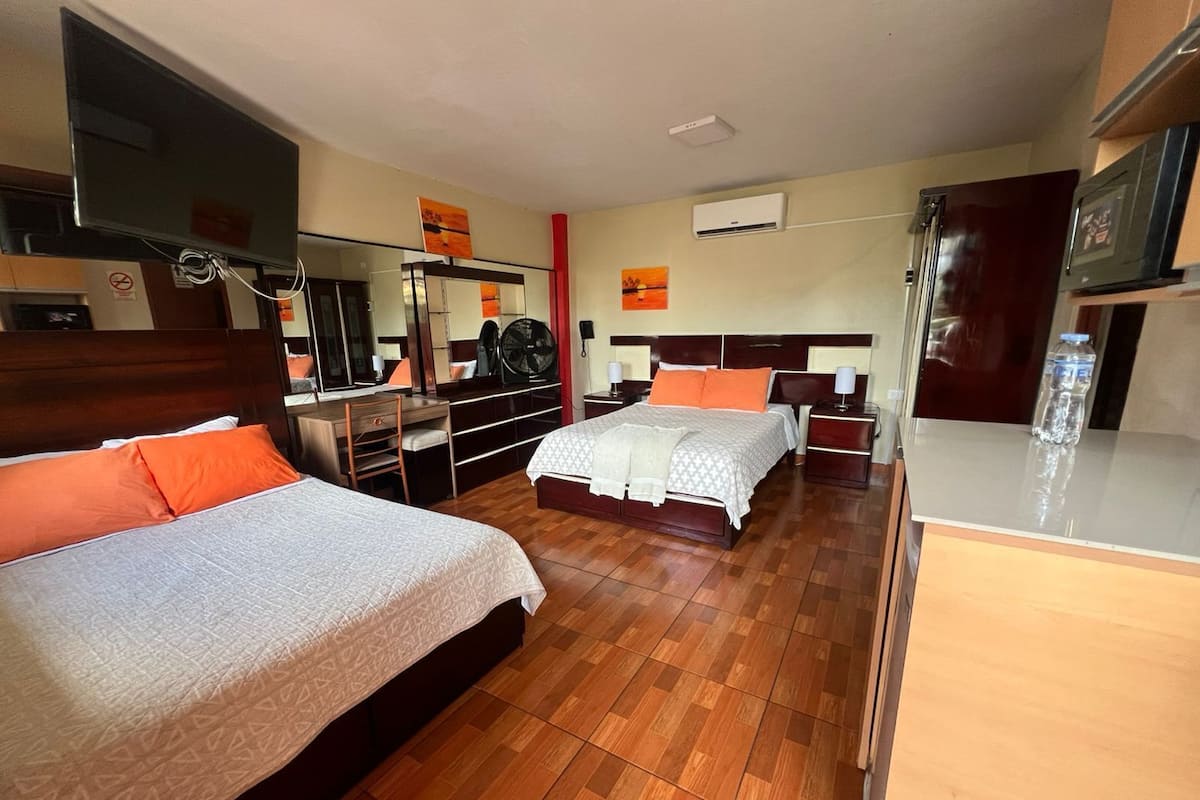 A hotel room with two beds, a desk, a chair, a TV, an air conditioning unit, and a water bottle on the counter.