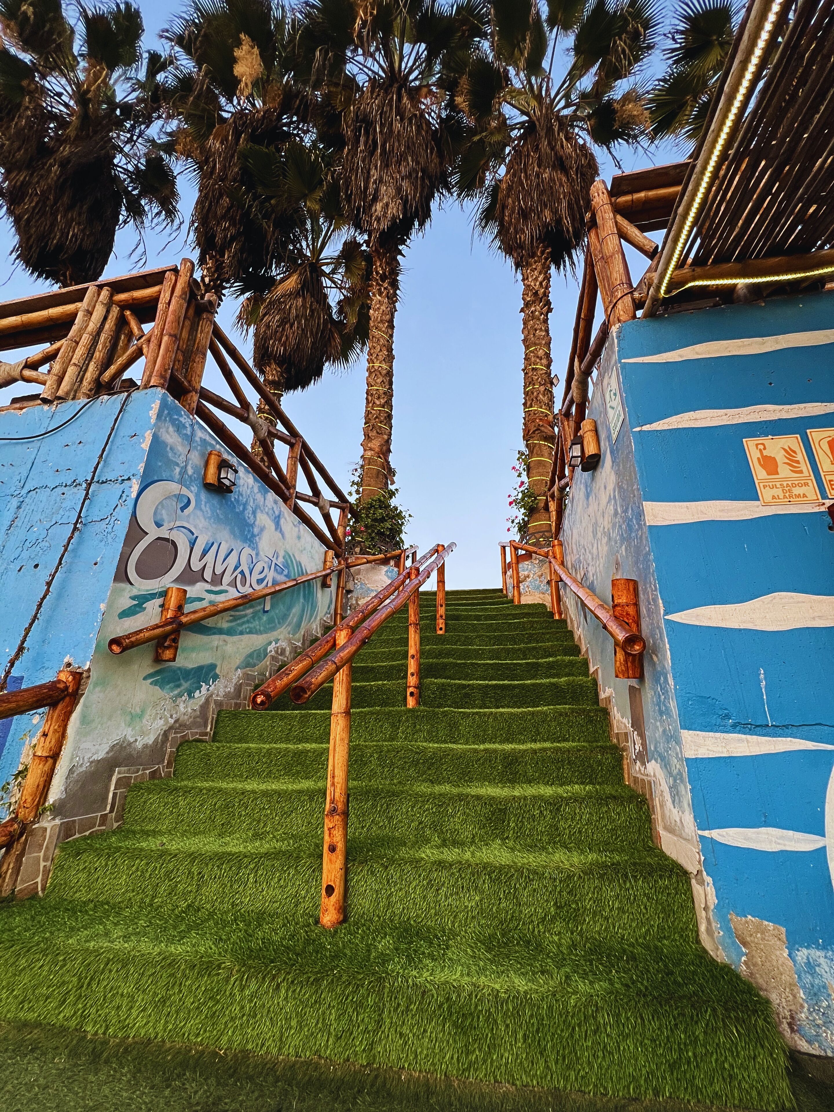 Una escalera con barandillas de madera y césped artificial, que conduce a un edificio de fachada azul y blanca.
