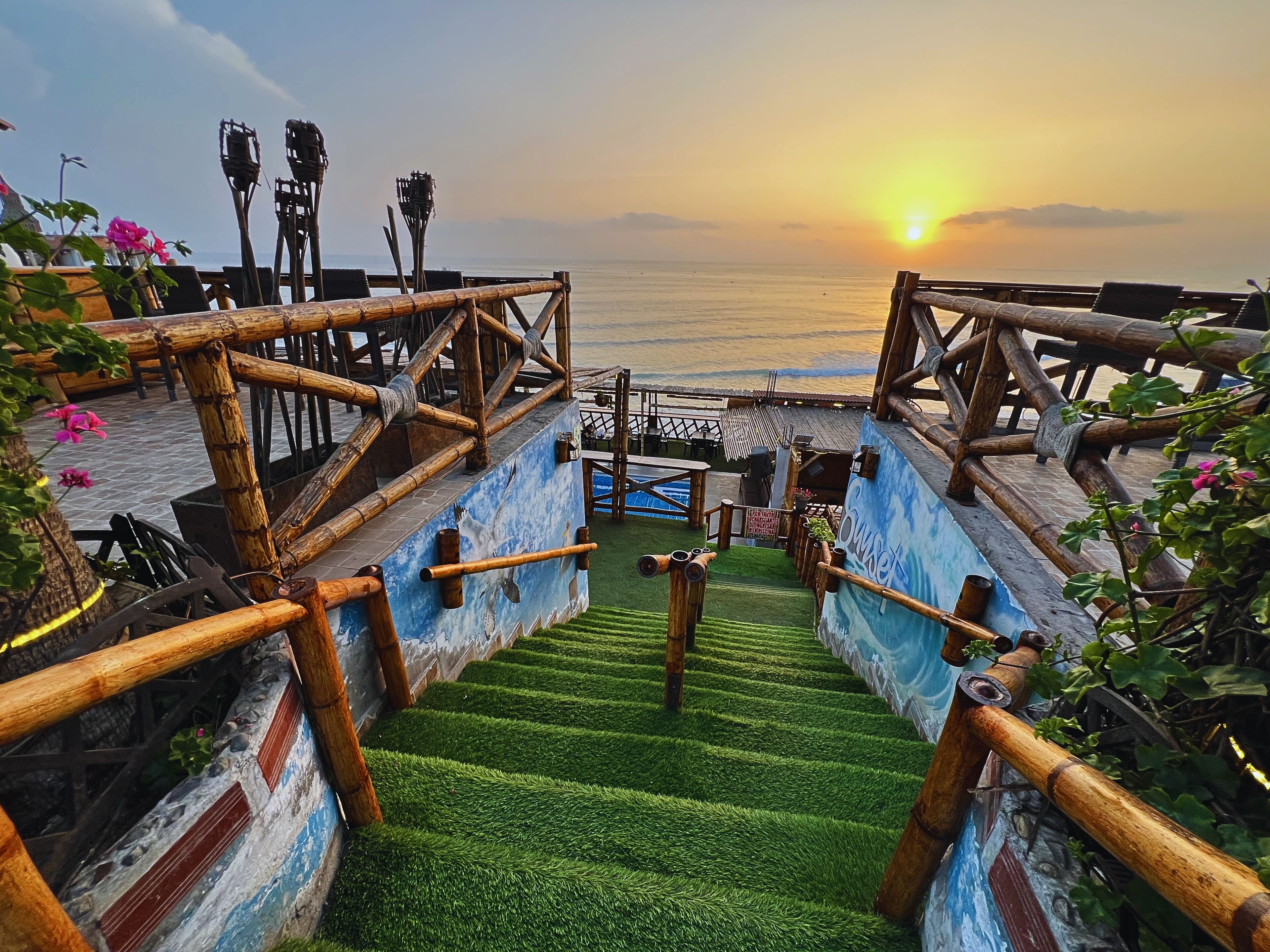 Uma escada de madeira leva a um deck com vista para o pôr do sol sobre o oceano.