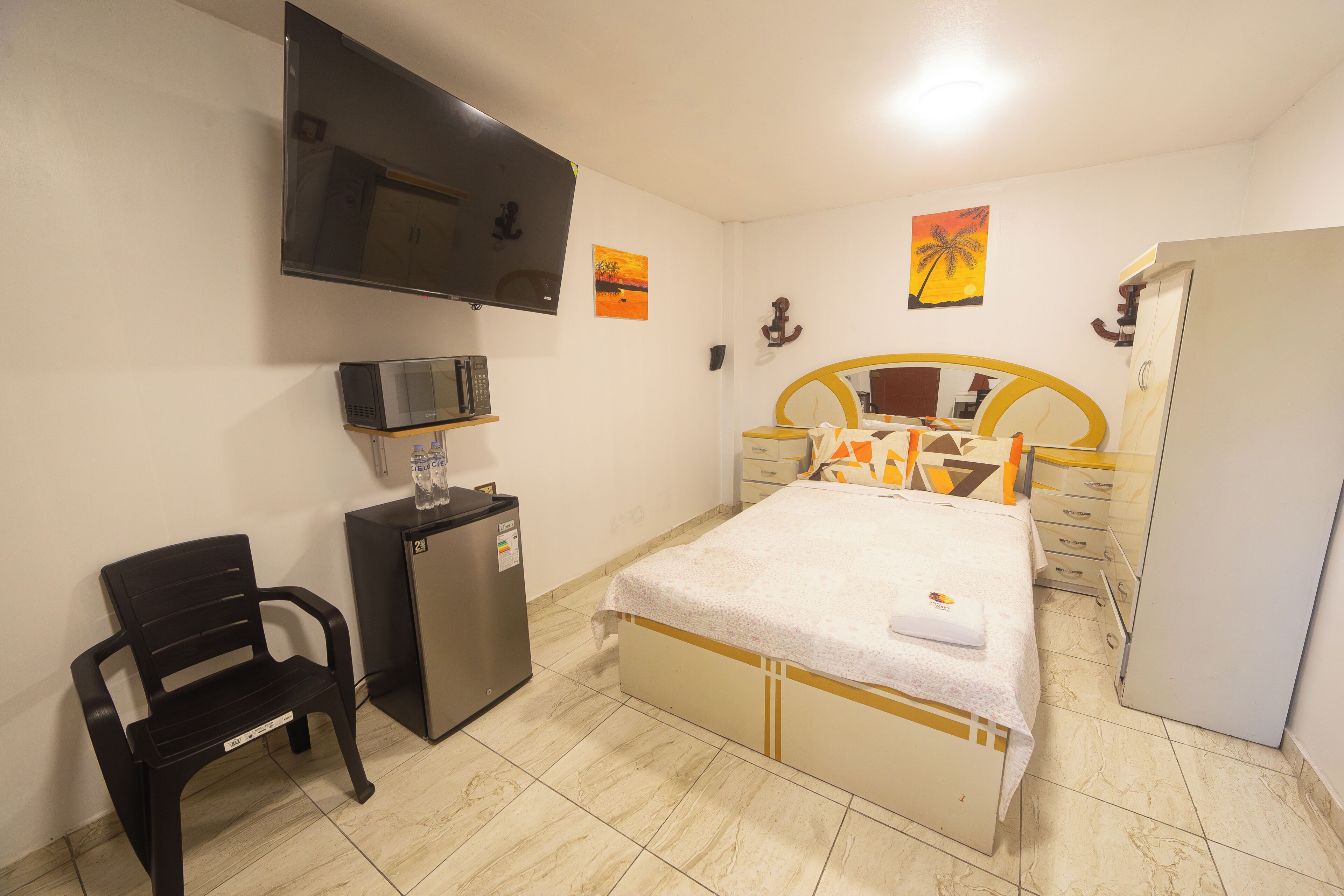A bedroom with a bed, a TV, a desk with a chair, a wardrobe, and artwork on the walls.