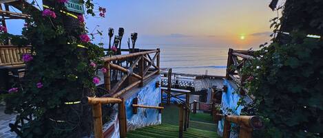 A staircase leading to a beach with a sunset view, surrounded by greenery and bamboo railings.