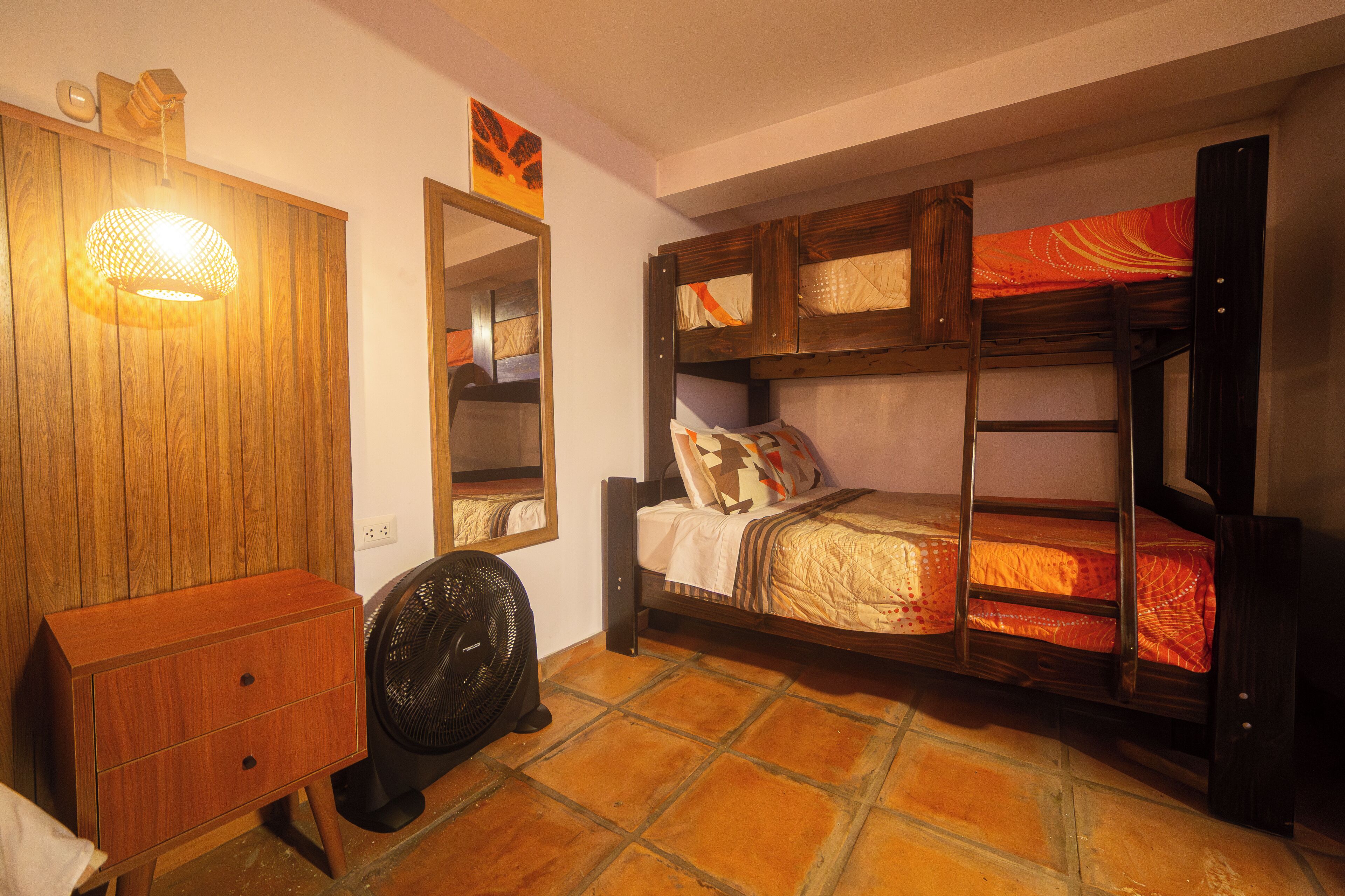 Une chambre avec un lit superposé, une commode en bois, un ventilateur et un miroir.