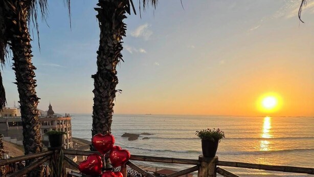 Une table à manger installée à l’extérieur, décorée de motifs en forme de cœur rouge, et offrant une vue sur le coucher de soleil sur la mer.