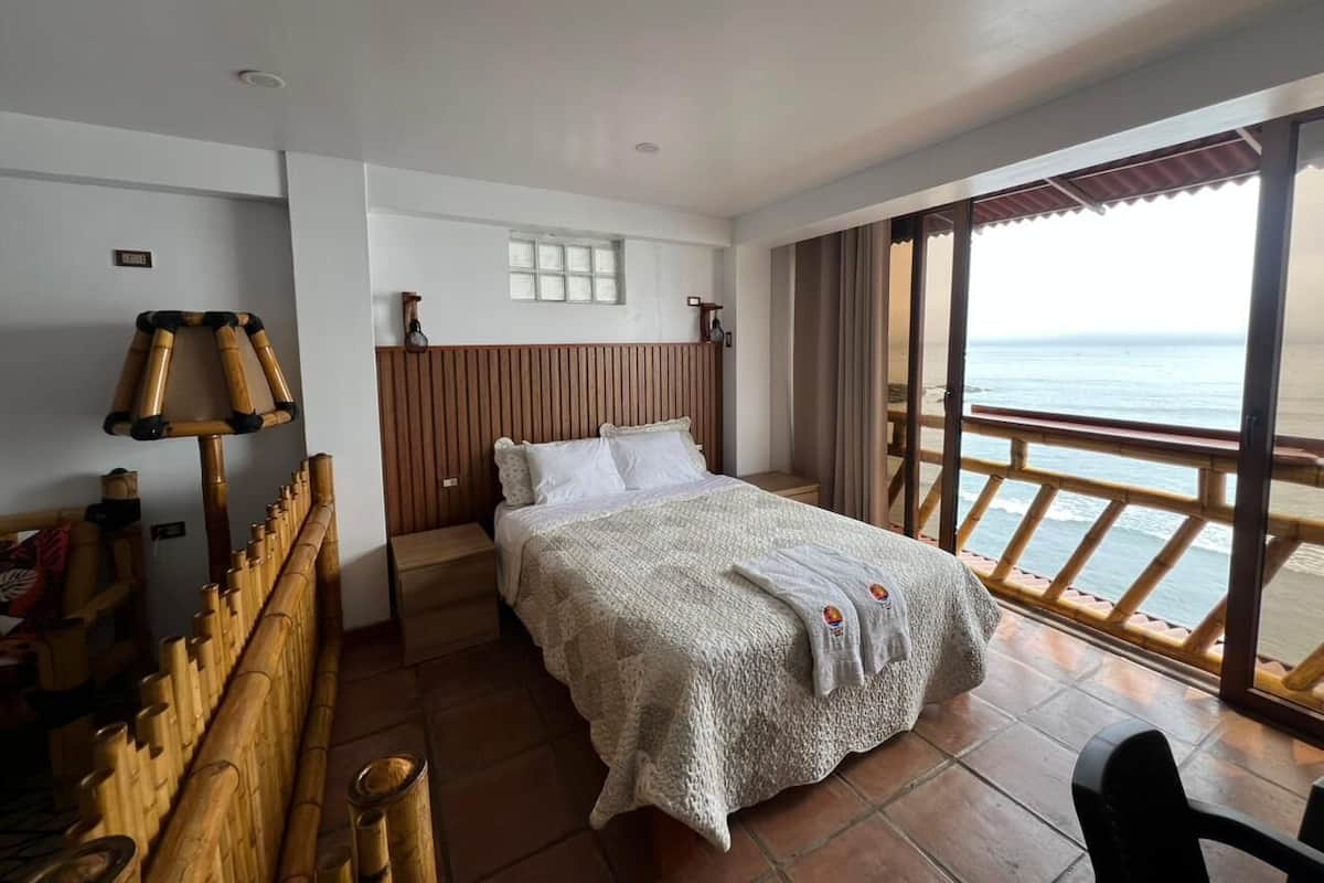A hotel room with a bed, a wooden headboard, a window with a view of the ocean, and a balcony with a railing.