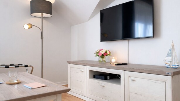 A modern living room with a flat-screen TV mounted on the wall, a wooden table with a glass of water, a vase with flowers, a candle, and a small model sailboat. - Welcome to “Strandmuschel” – the largest of our cozy one-room apartments. (Ostseebad Binz)
