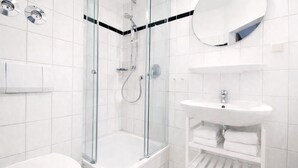 A modern bathroom with a glass-enclosed shower, a white sink, and a mirror. - Welcome to “Strandmuschel” – the largest of our cozy one-room apartments. (Ostseebad Binz)