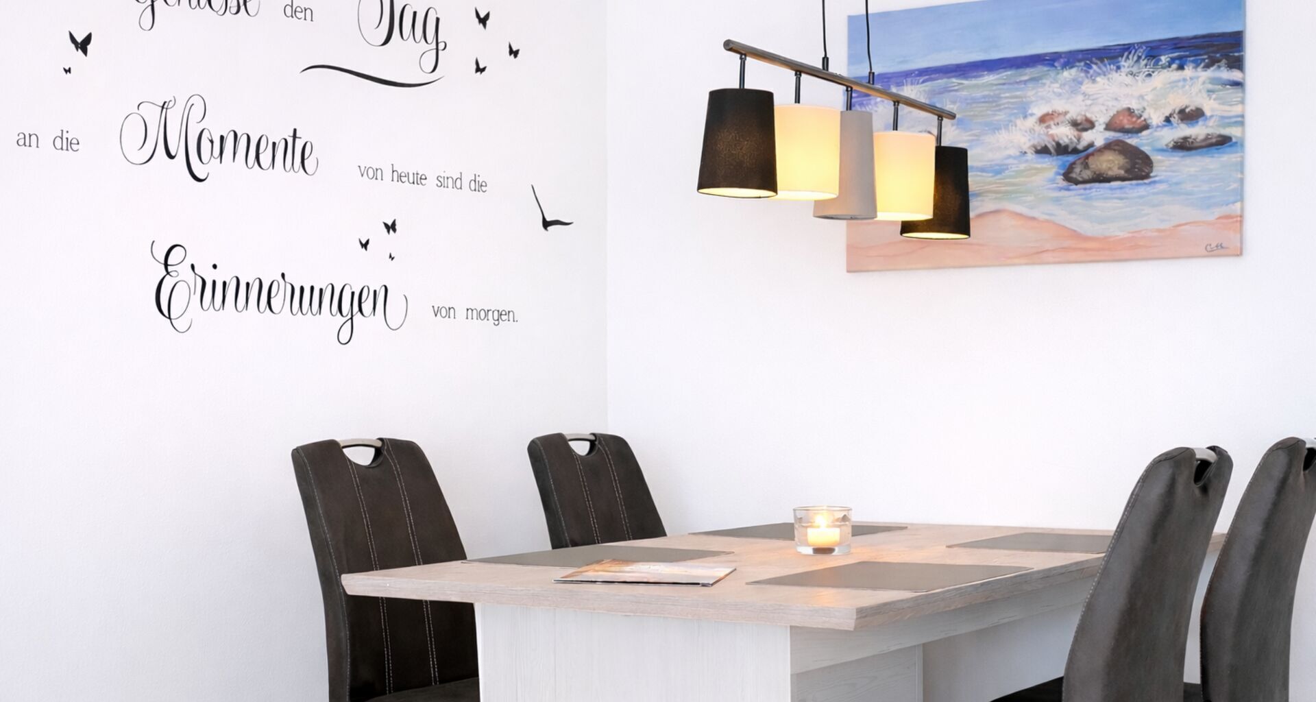 Een eethoek met een tafel, stoelen, een kaars, en een muur met een schilderij en tekst.