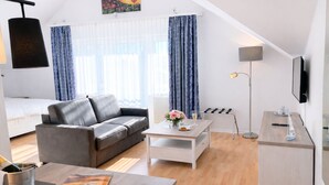 A modern, well-lit room with a grey sofa, a small white table, a bed, and a TV mounted on the wall. - Welcome to “Strandmuschel” – the largest of our cozy one-room apartments. (Ostseebad Binz)