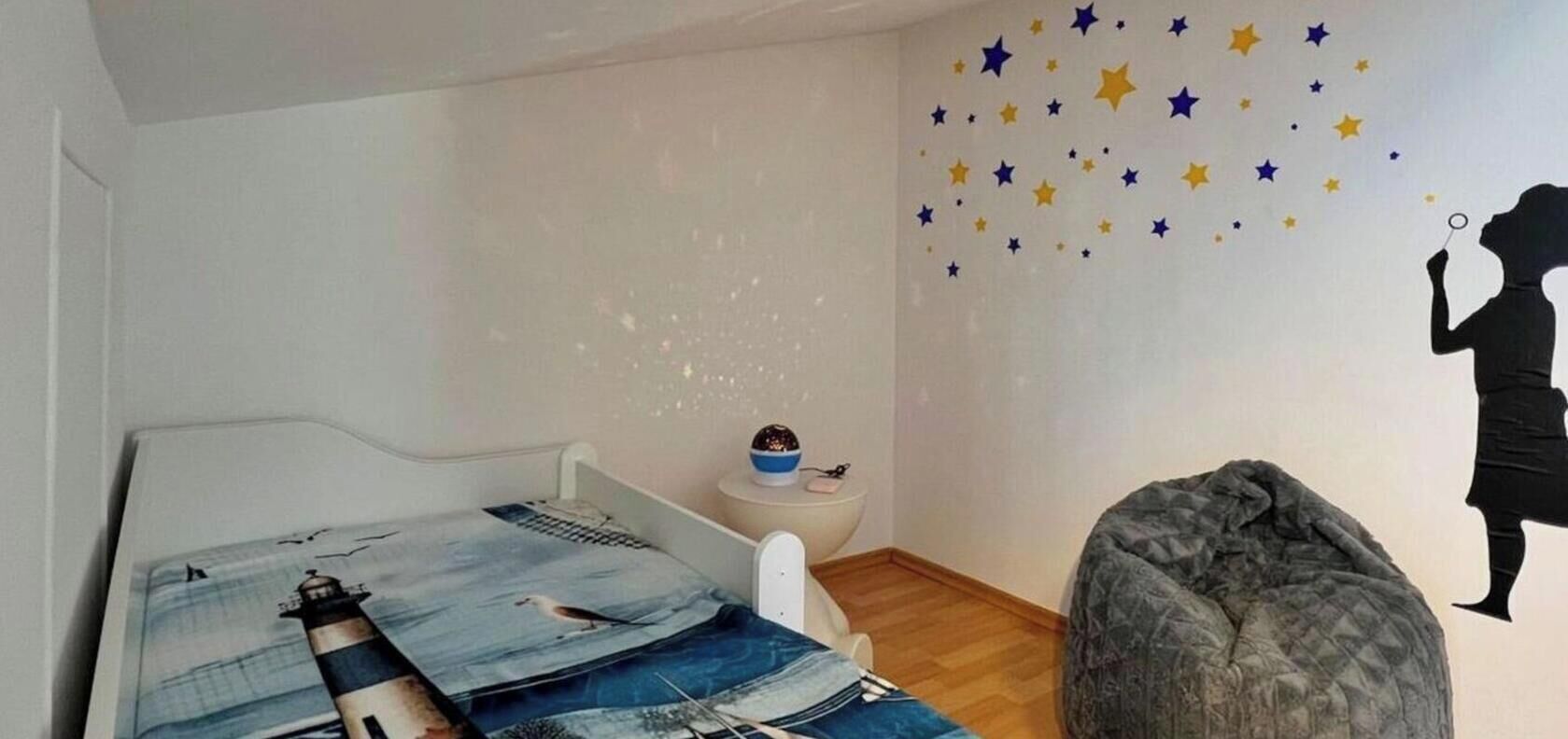 Een kinderkamer met een bed, een klein krukje, een kruikzak en een muur met sterstickers.