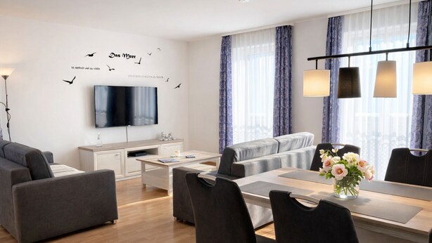 Een moderne woonkamer met een flatscreen-televisie, grijze banken, een eettafel met stoelen en een vaas met bloemen.