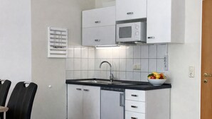 Een moderne keuken met witte kasten, een RVS vaatwasser, een magnetron en een spoelbak. Er staat een fruitschaal op het aanrecht en een raam met jaloezieën.